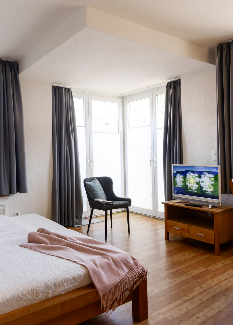 Blick vom Zimmer über das Doppelbett zur Terrassentür. Vor dem hellen Fenster steht ein moderner Stuhl mit Kissen. Rechts daneben steht ein Flachbildfernseher auf einer TV-Kommode.