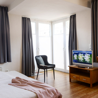 Gästewohnung der TRAVE "Auf dem Sande" Blick vom Zimmer über das Doppelbett zur Terrassentür. Vor dem hellen Fenster steht ein moderner Stuhl mit Kissen. Rechts daneben steht ein Flachbildfernseher auf einer TV-Kommode.