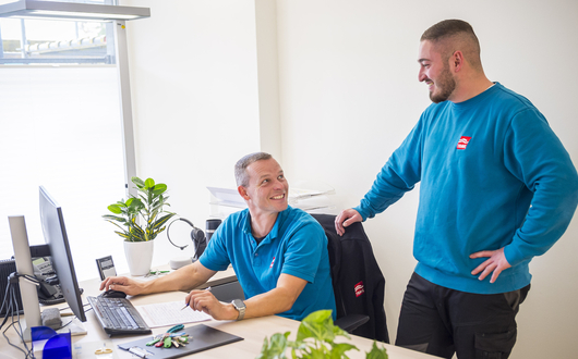 Hauswarte in den Quartieren Zwei Hauswarte der TRAVE. Einer sitzt am Schreibtisch vor dem Computer, der andere steht hinter im. Beide schauen sich an und lächeln.