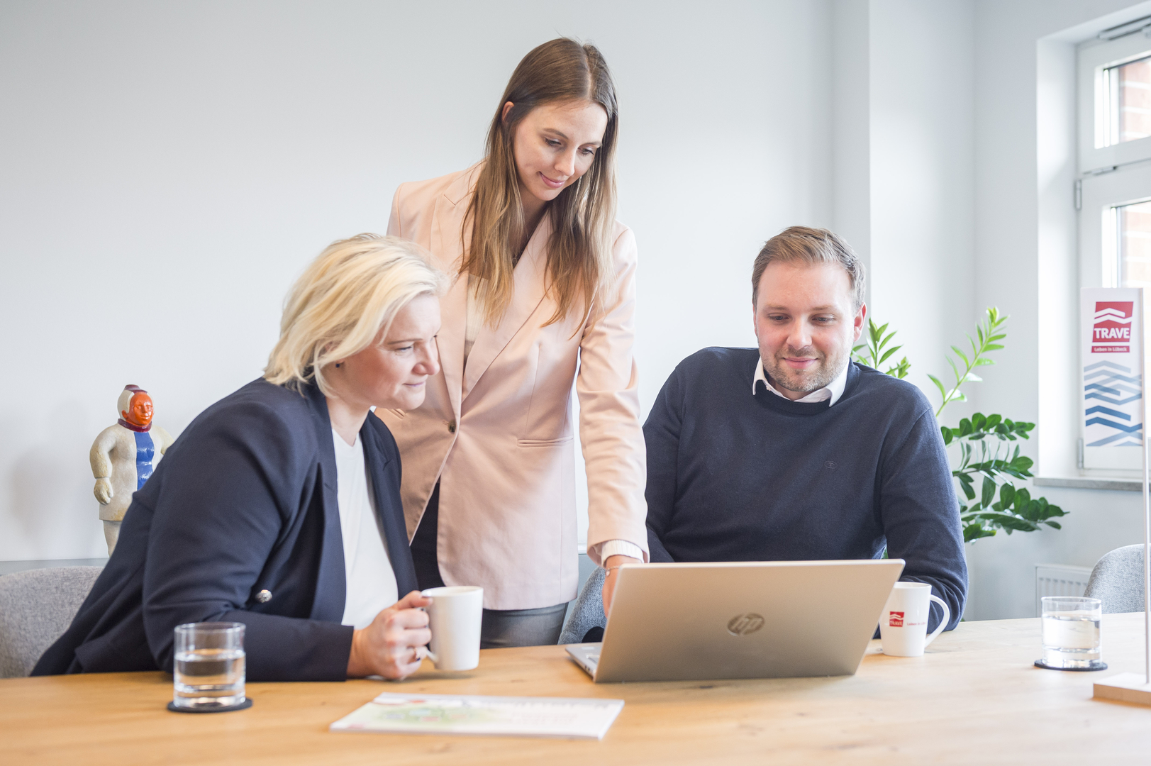 Drei Personen sitzen gemeinsam an einem Tisch und arbeiten an einem Laptop in einem hellen Büro.